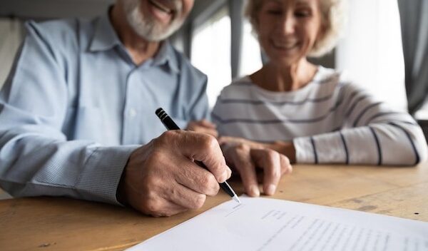Close up focus on wrinkled male hand signing paper document Smiling elderly mature family couple putting signature on leasing or medical insurance contract purchase agreement with real estate agent