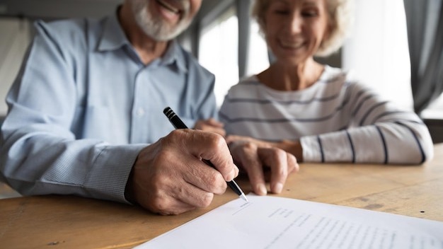 Close up focus on wrinkled male hand signing paper document Smiling elderly mature family couple putting signature on leasing or medical insurance contract purchase agreement with real estate agent