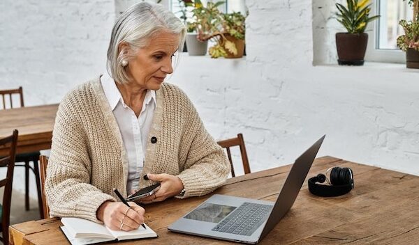 Mature woman with gray hair engaged in online work at a cozy wooden table in a bright space