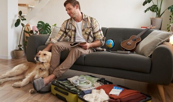 Smiling handsome guy and his dog sitting in living room packing suitcase for summer holiday planning