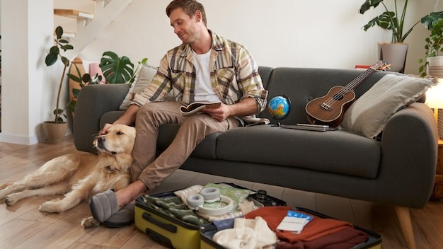 Smiling handsome guy and his dog sitting in living room packing suitcase for summer holiday planning