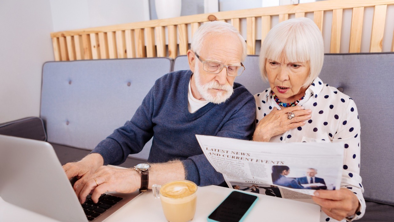 Breaking news. Amazed senior couple examining newspaper and sitting at table