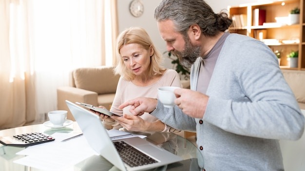 Mature bearded man with cup of tea pointing at document held by his wife while both looking it through