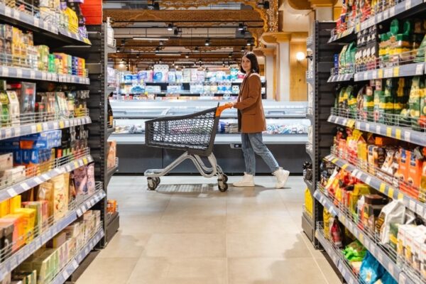 A woman with a cart walks between rows of shelves in a grocery store
