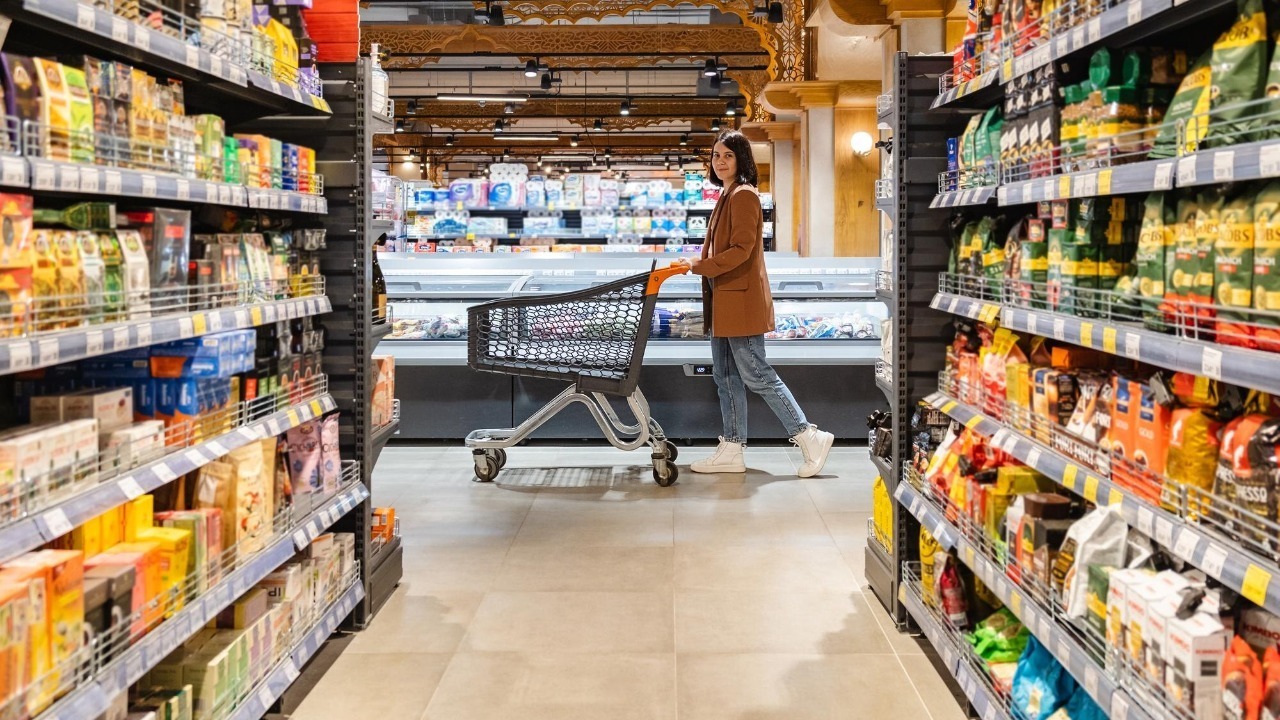 A woman with a cart walks between rows of shelves in a grocery store