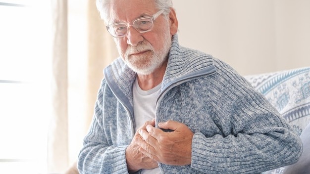 Worried 70s senior man in light blue sweater sitting at home on sofa suffering from bad chest pain elderly heart disease