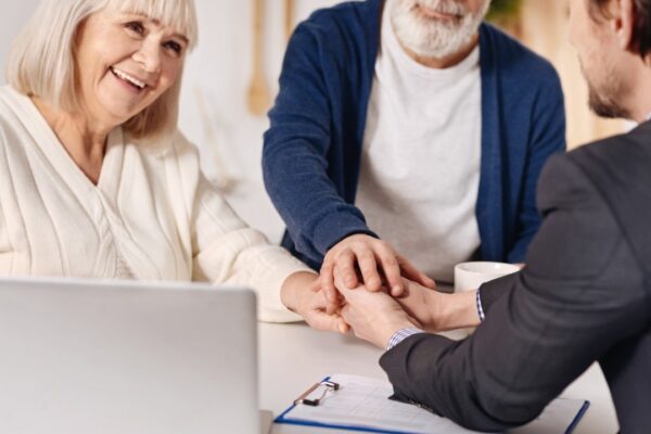Full of joy. Smiling delighted old couple sitting at home and concluding agreement with real estate agent while shaking hands and expressing happiness