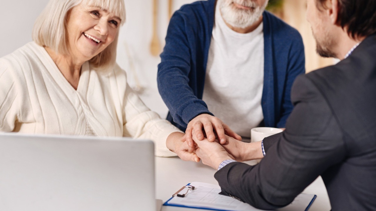 Full of joy. Smiling delighted old couple sitting at home and concluding agreement with real estate agent while shaking hands and expressing happiness