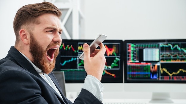 Angry irritated bearded young businessman screaming in smartphone in office