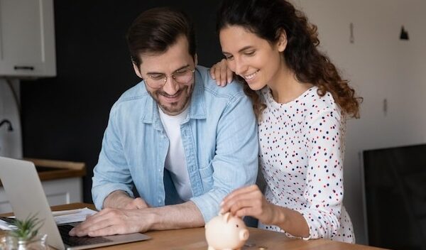 Young married couple in domestic kitchen with laptop put coins in piggy bank spouses saving money planning family budget making investment in future down payment on first affordable house concept