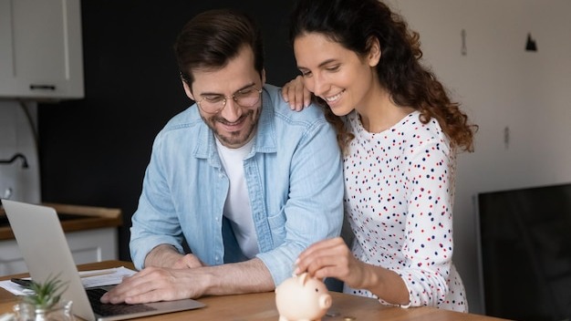 Young married couple in domestic kitchen with laptop put coins in piggy bank spouses saving money planning family budget making investment in future down payment on first affordable house concept