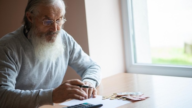 Old bearded senior man with calculator and bills counting euro money and writing notes