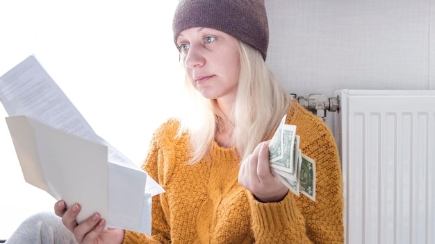 A young girl in a yellow sweater and a brown hat is sitting on the floor counting money and thinking how to pay bills and taxes near a heater with a thermostat