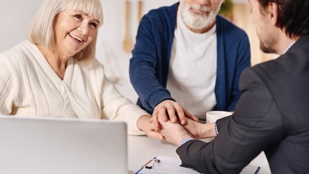 Full of joy. Smiling delighted old couple sitting at home and concluding agreement with real estate agent while shaking hands and expressing happiness