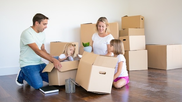 Mom, dad and little daughters unpacking things in new apartment, sitting on floor and taking objects from open boxes