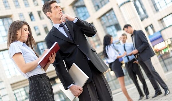 Young businessman and his secretary standing in front of office building. Businessman carrying in one hand a closed laptop and thinking, and the secretary keeps the planner.