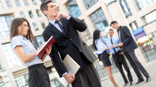 Young businessman and his secretary standing in front of office building. Businessman carrying in one hand a closed laptop and thinking, and the secretary keeps the planner.
