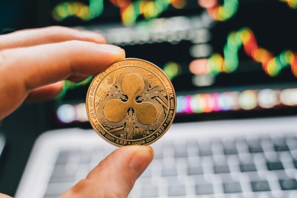 A close-up view of a hand holding a Ripple cryptocurrency coin with a blurred computer in the background.