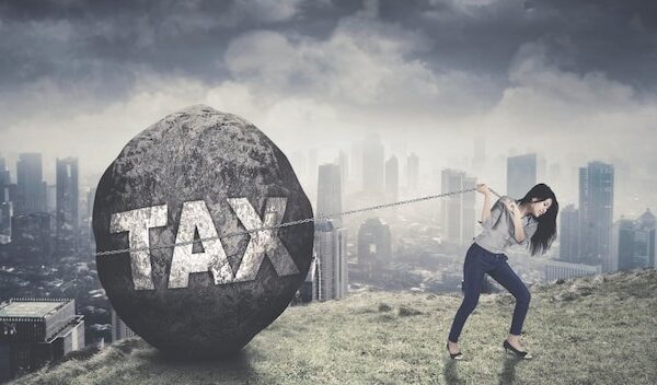 Asian female entrepreneur struggling to pull tax word on a stone while climbing on the mountain with modern city background
