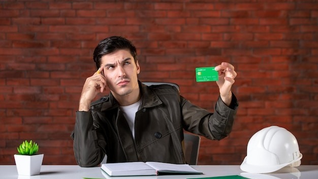 Front view male engineer sitting behind his working place and holding green credit card builder money plan corporate contractor business property