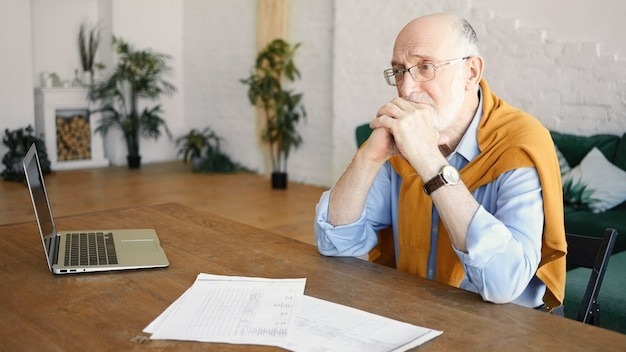 Indoor shot of unhappy sad senior bearded male entrepreneur sitting at desk with laptop and papers having depressed facial expression, frustrated with financial problems, holding hands under his chin