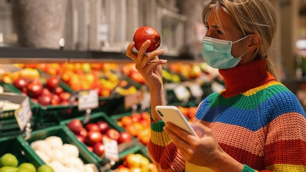 Woman at supermarket in the fruit section choosing fruit with smartphone wearing an anti virus protection mask due to covid19 Coronavirus pandemic