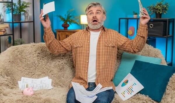 Mature man on sofa using smartphone calculator counting cash and bills feeling exhausted at home