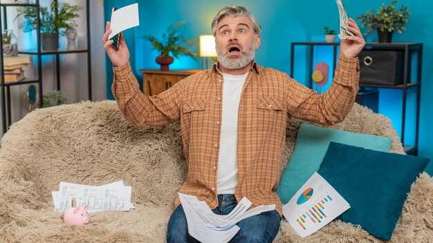 Mature man on sofa using smartphone calculator counting cash and bills feeling exhausted at home