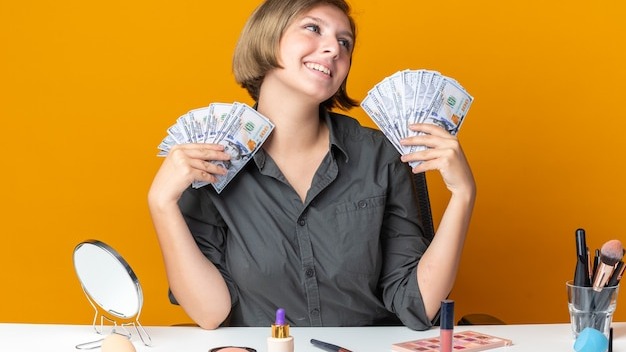 Smiling tilting head beautiful woman sits at table with makeup tools holding cash