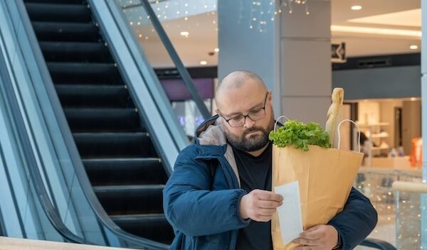 Surprised overweight man with shopping paper bag full of food looks at paper receipt on escalator in