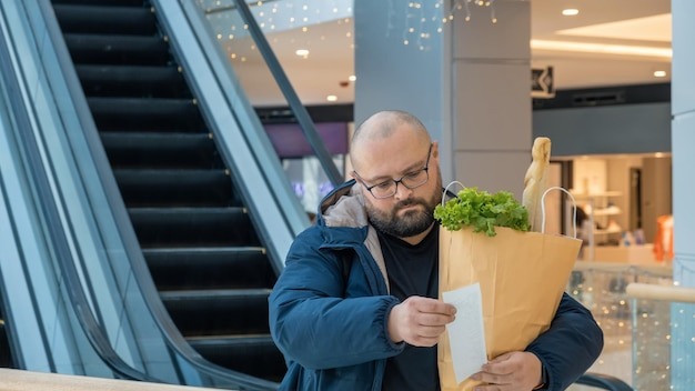 Surprised overweight man with shopping paper bag full of food looks at paper receipt on escalator in