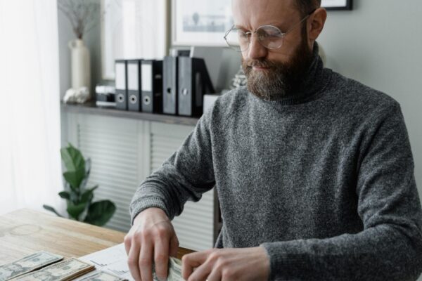 An adult man in a gray sweater counting dollar bills at a wooden desk, showcasing financial management.