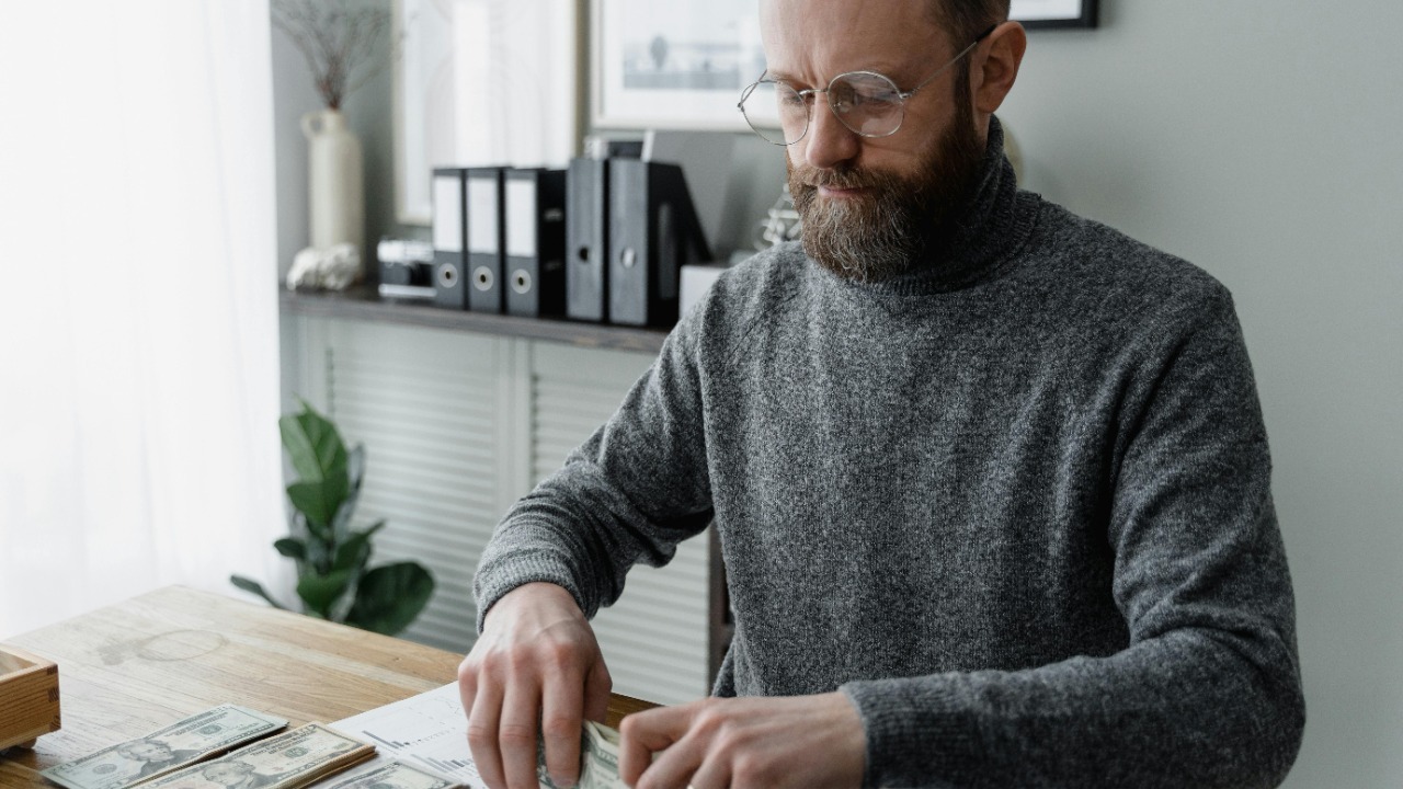 An adult man in a gray sweater counting dollar bills at a wooden desk, showcasing financial management.