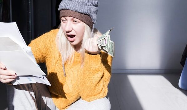 A young girl in a yellow sweater and two hats is horrified to study large bills taxes and utility bills