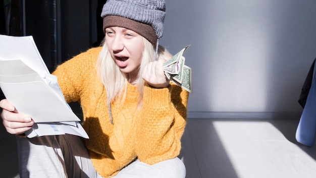 A young girl in a yellow sweater and two hats is horrified to study large bills taxes and utility bills
