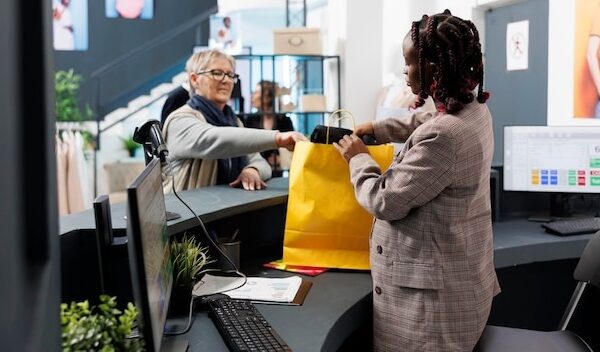 African american showroom worker preparing purchase for elderly client in clothing store. Shopaholic woman buying fashionable casual wear, making electronic payment at pos terminal.