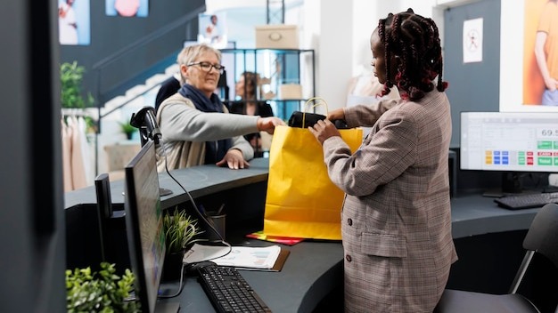 African american showroom worker preparing purchase for elderly client in clothing store. Shopaholic woman buying fashionable casual wear, making electronic payment at pos terminal.