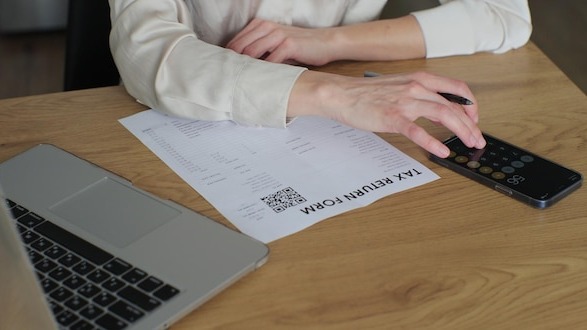 Young woman is writing tax return form and looking at laptop at table in home room Close up woman fills out paper document and counts on the phone screen