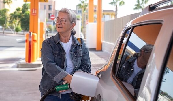 Mature modern elderly woman fills up her car at selfservice fuel pump in European gas station inflation price increase economy speculation concept