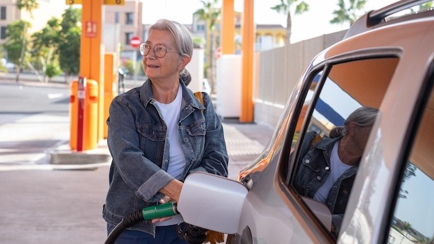 Mature modern elderly woman fills up her car at selfservice fuel pump in European gas station inflation price increase economy speculation concept