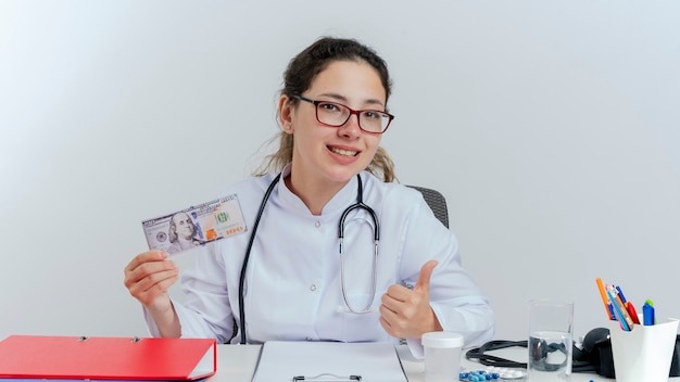 Happy young female doctor wearing medical robe and stethoscope and glasses sitting at desk with medical tools holding money looking showing thumb up isolated