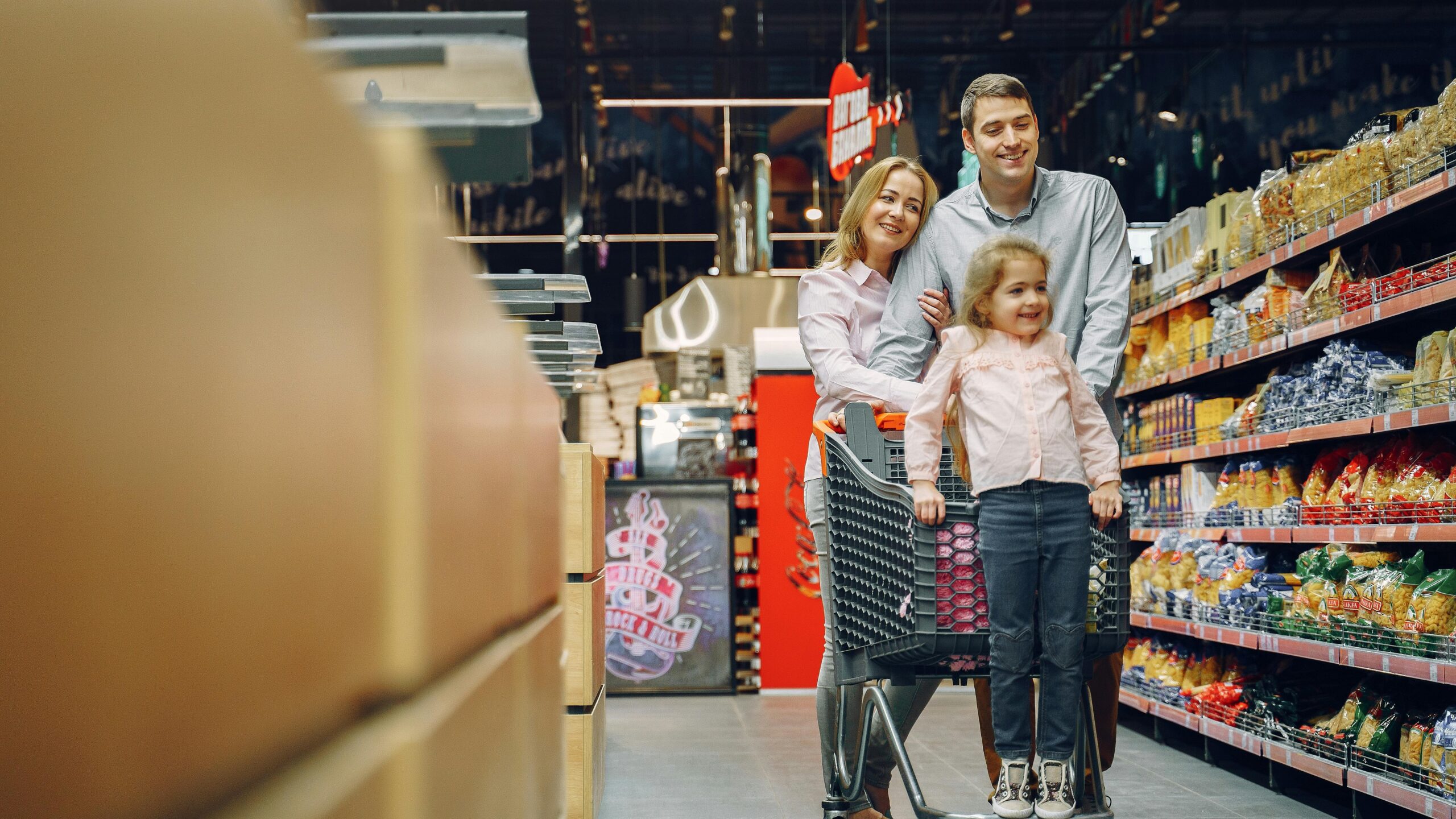 Family enjoying a shopping trip in a supermarket, with a child in a cart and parents smiling.