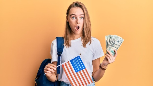 Beautiful blonde woman exchange student holding usa flag and dollars banknotes in shock face, looking skeptical and sarcastic, surprised with open mouth