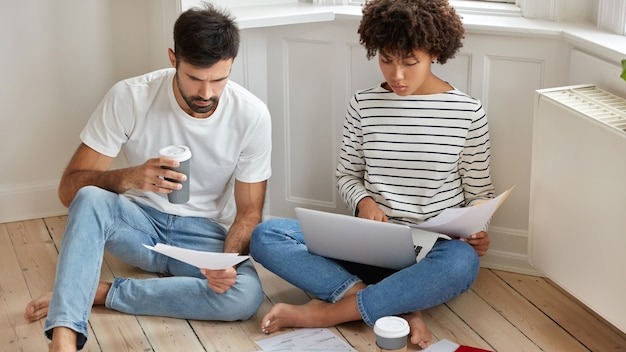 People, business and work concept. Woman and man coworkers study documentation and think about productive strategy to raise profits, pose on wooden floor with takeaway coffee, have serious look