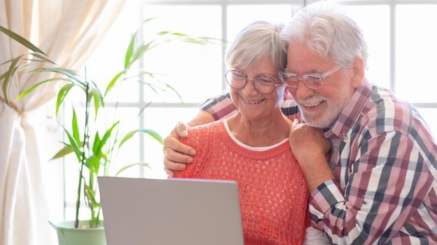 Beautiful caucasian senior couple with eyeglasses browsing together on laptop sitting at home old retirees people enjoying tech and social