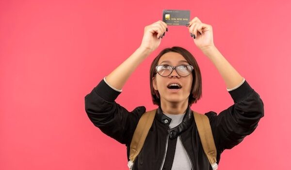 Impressed young student girl wearing glasses and back bag holding and looking at credit card isolated on pink