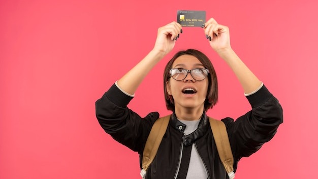 Impressed young student girl wearing glasses and back bag holding and looking at credit card isolated on pink