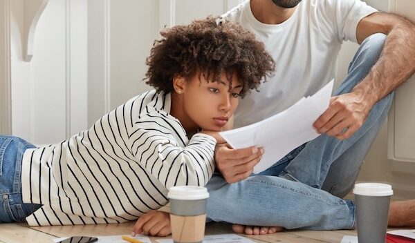 Female and male managers pose at floor, discuss report, learns sales result from papers, meet at domestic atmosphere, drinks coffee from disposable cup, analyze company profit. Collaboration concept