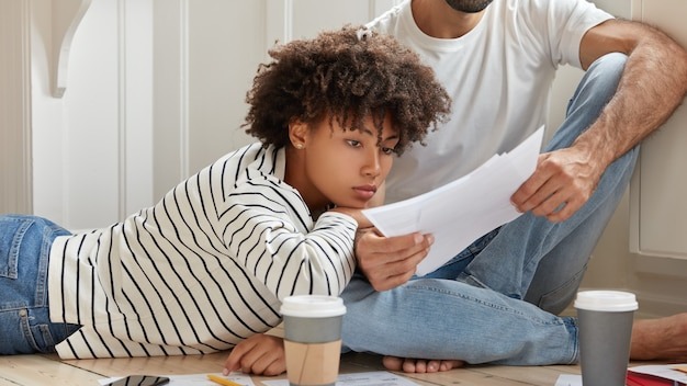 Female and male managers pose at floor, discuss report, learns sales result from papers, meet at domestic atmosphere, drinks coffee from disposable cup, analyze company profit. Collaboration concept