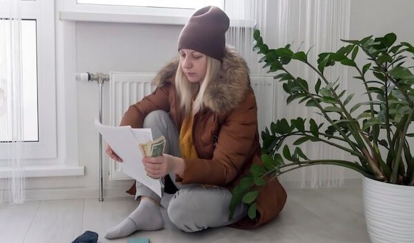 A woman warmly dressed in a brown jacket and hat is sitting near a heating appliance with bills in her hands money and a phone The concept of a crisis and a large set for utilities